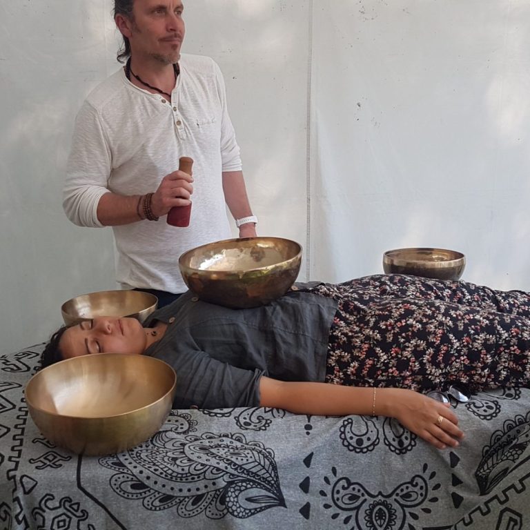 Soin et massage sonore individuel aux bols tibétains Une personne allongée reçoit un soin sonore avec plusieurs bols tibétains placés sur son corps, guidé par un praticien dans un espace calme.