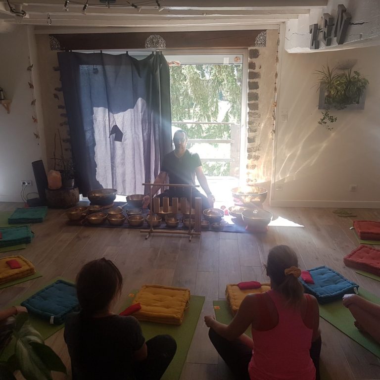 Cercle de méditation sonore au bol tibétain Un groupe de personnes assises en cercle sur des tapis, écoutant un praticien jouant des bols tibétains dans une salle lumineuse.